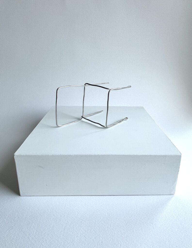 Small aluminium wire chair lying on its side on a white panel, seen from the front