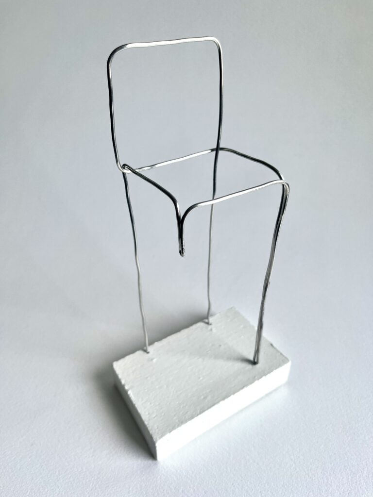 Small aluminium wire chair with one shortened leg on a white panel, seen from above