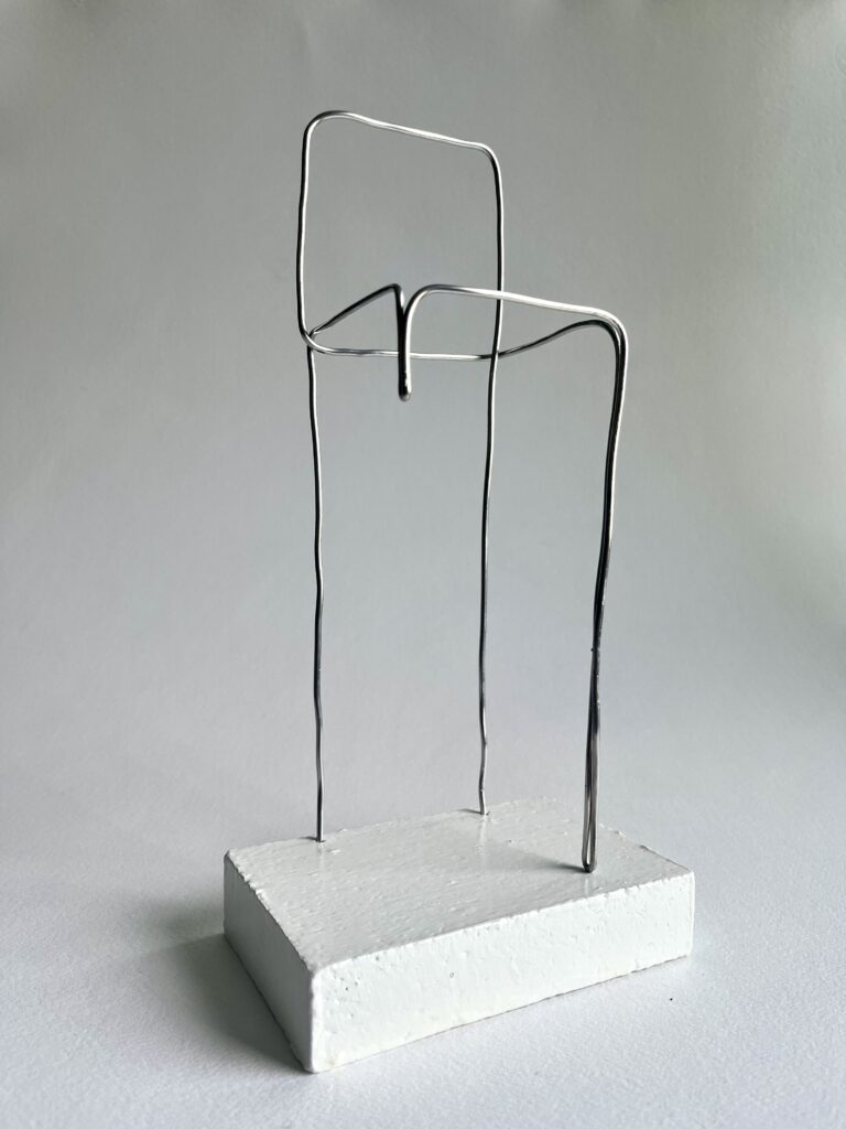 Small aluminium wire chair with one shortened leg on a white panel, seen from below