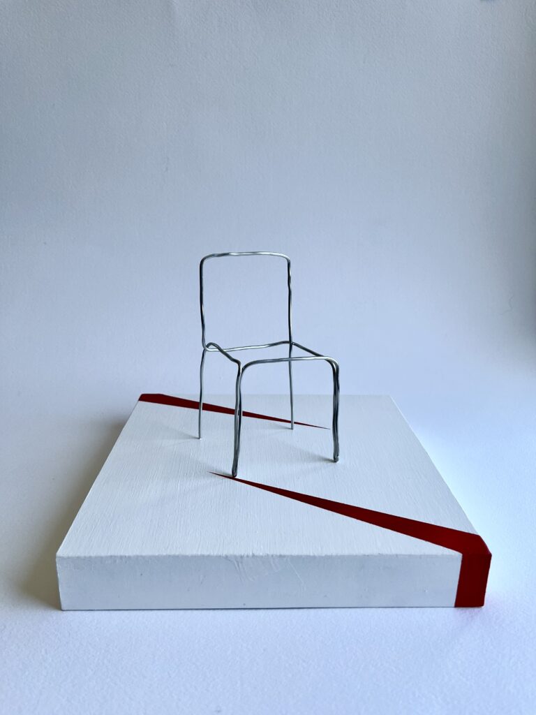 Small aluminium wire chair standing upright on a white panel, seen from the front