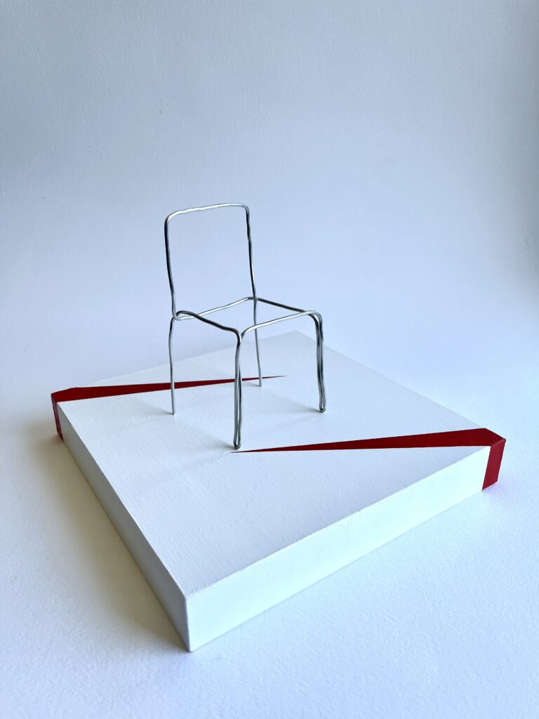 Small aluminium wire chair standing upright on a white panel, angled view