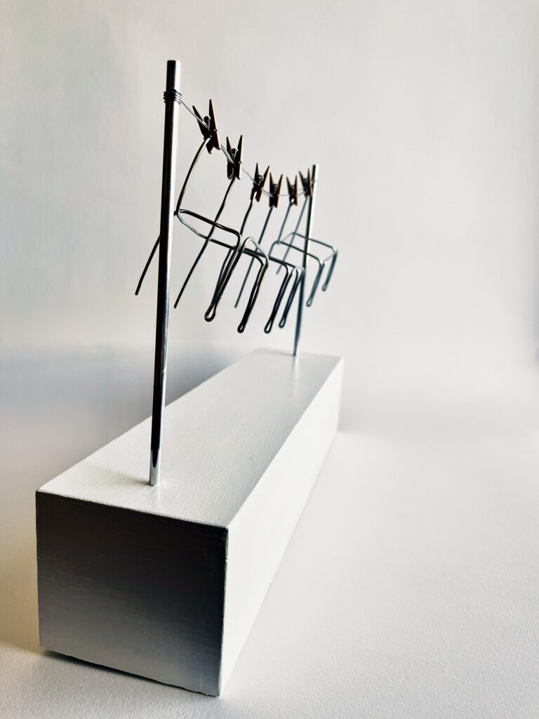 Three small aluminium wire chairs pegged with wooden clothespins to a wire line strung between two aluminium poles on a white plinth, angled view