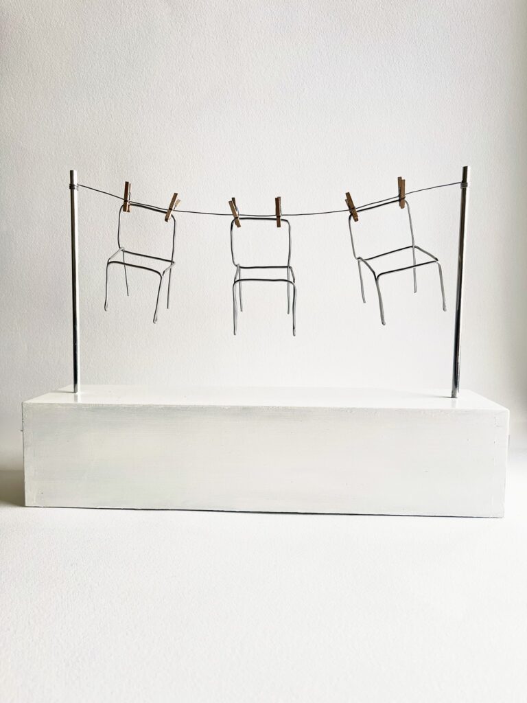 Three small aluminium wire chairs pegged with wooden clothespins to a wire line strung between two aluminium poles on a white plinth, seen from the front